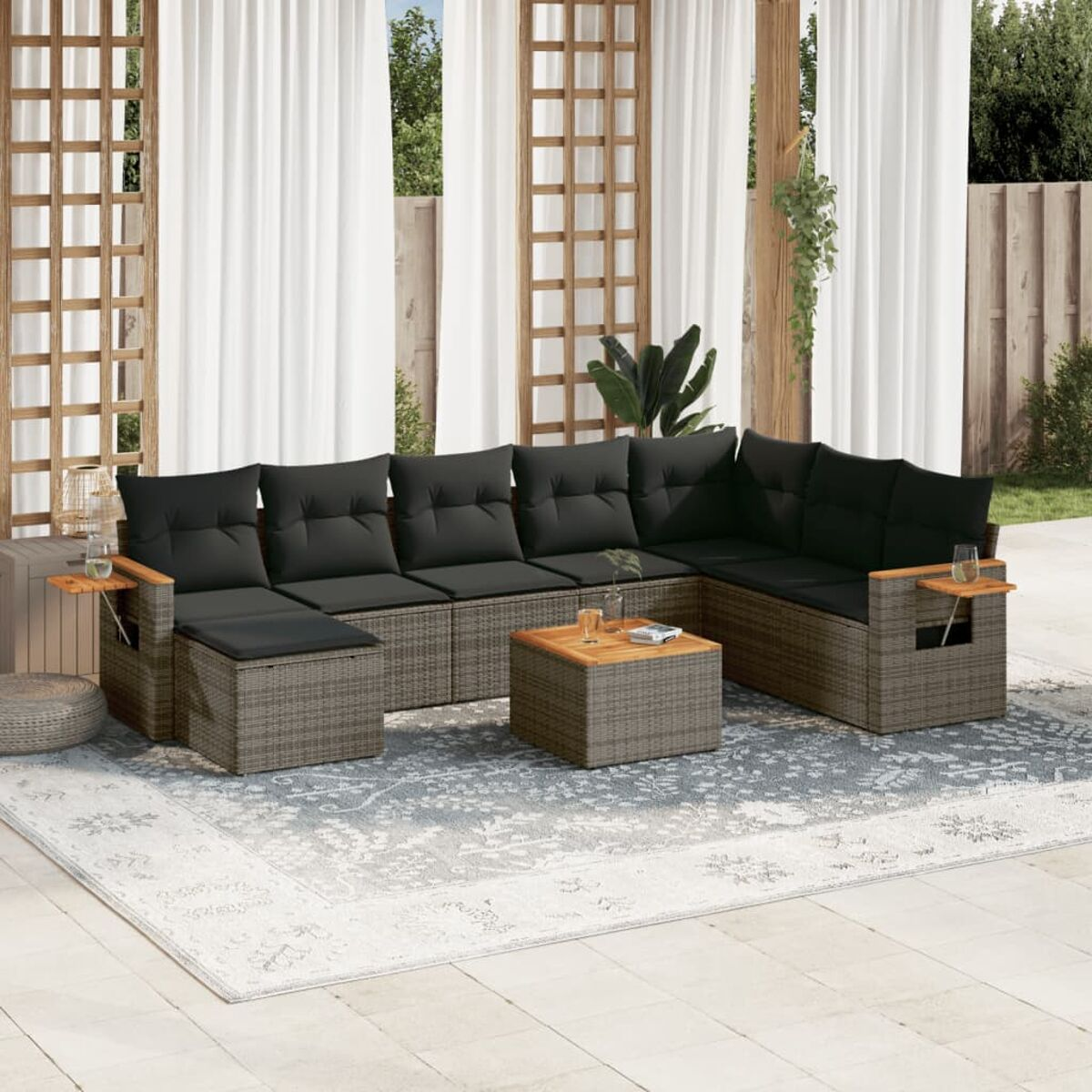 Graues Rattan-Ecksofa mit schwarzen Kissen, Holztisch und Teppich. Vorhänge und Grün im Hintergrund.