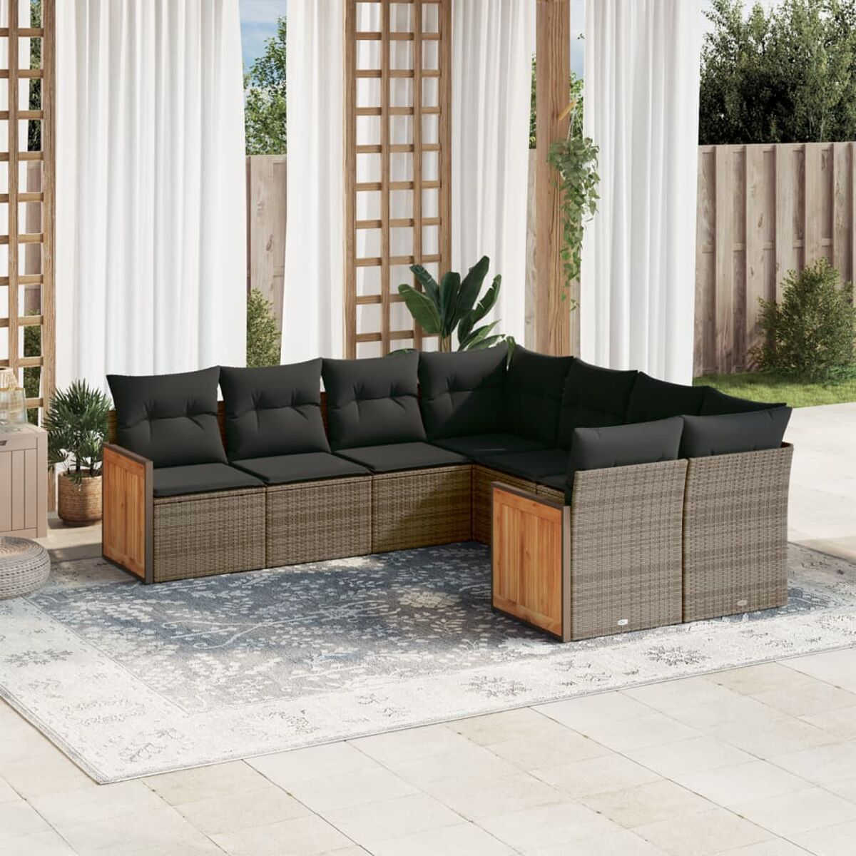 Ein graues Korbsitzmöbel-Ecksofa für den Außenbereich mit schwarzen Kissen. Es steht auf einer Terrasse mit Teppich, Vorhängen und Pflanzen.