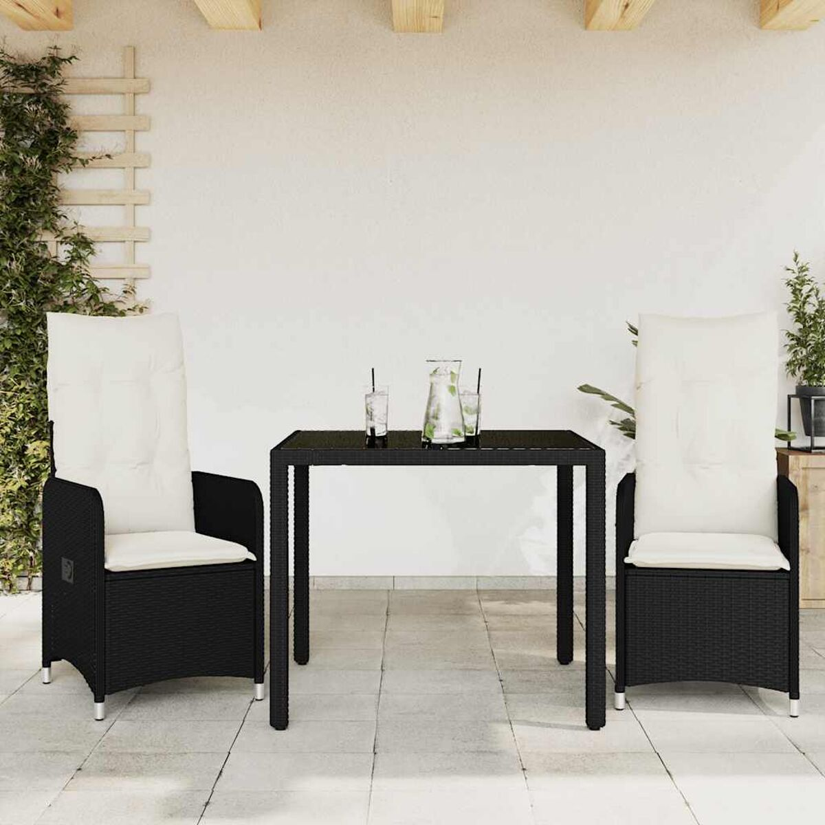 Tavolo nero con due sedie alte con cuscini bianchi. Ambiente esterno con muro bianco e piante.