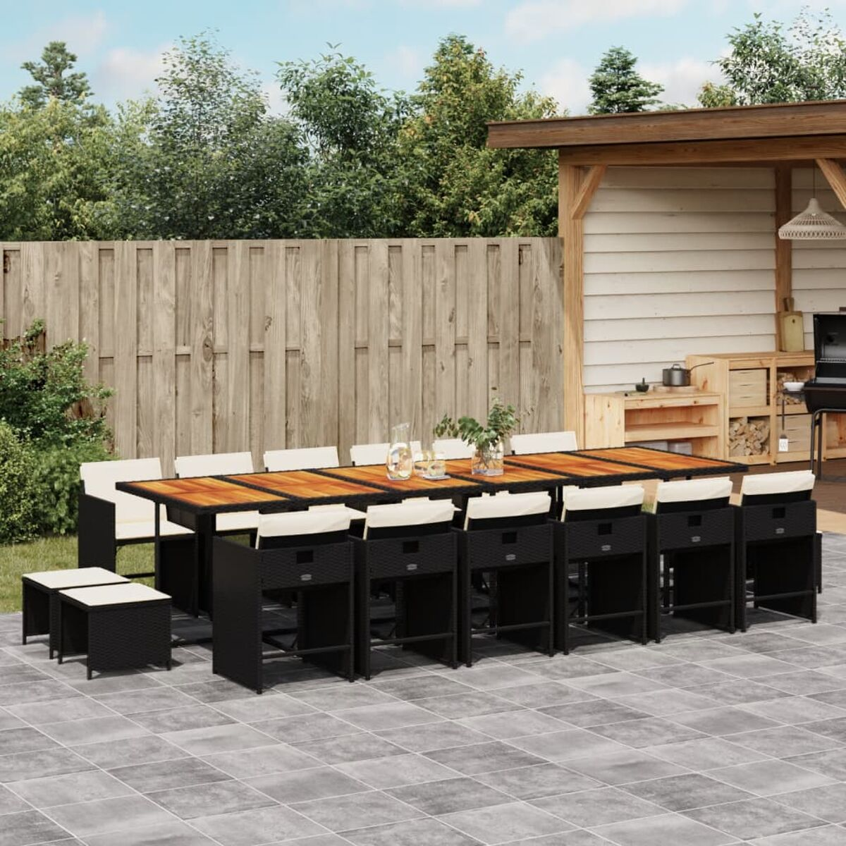 Essgruppe im Freien mit einem langen Tisch, Stühlen und Hockern auf einer Terrasse. Grüne Bäume im Hintergrund.
