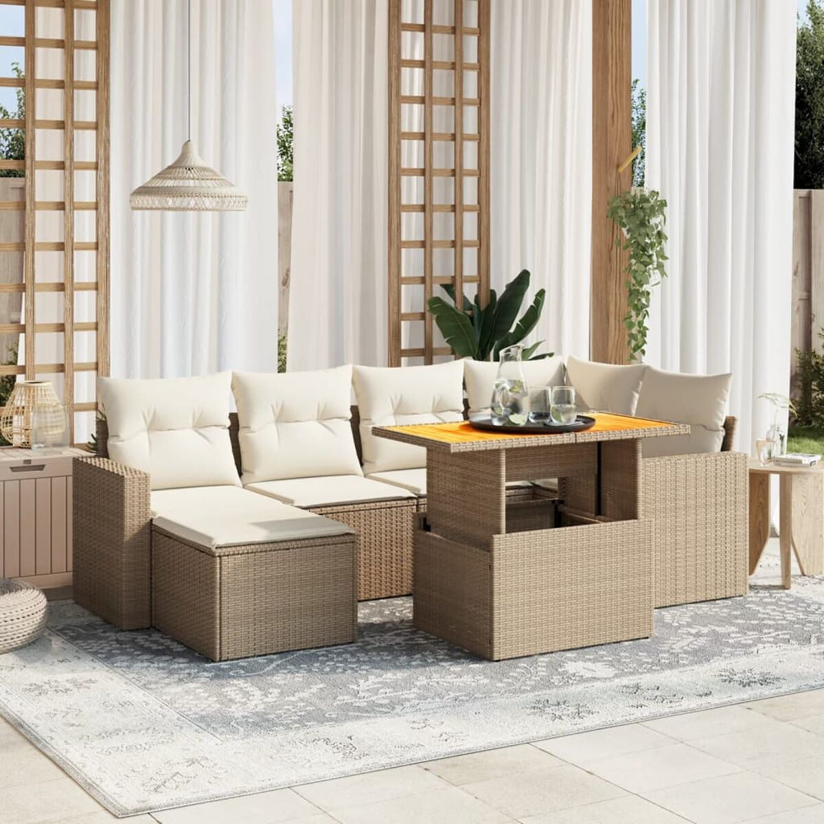 Set patio esterno con cuscini beige, struttura in vimini e tavolo sollevabile. Tende bianche e piante.