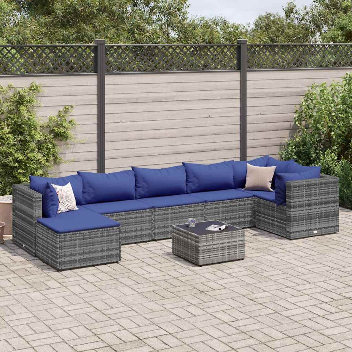 Graues Rattan-Ecksofa mit blauen Kissen, einem kleinen Tisch und Gartenhintergrund.