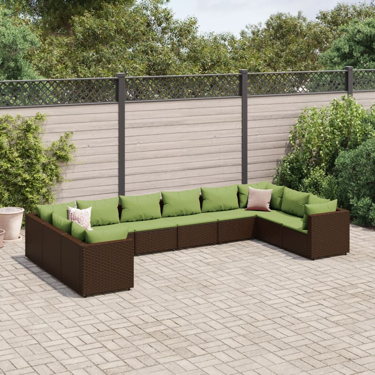 Braunes Outdoor-Ecksofa aus Korbgeflecht mit grünen Kissen. Auf Ziegelpflaster, grüne Blätter im Hintergrund.