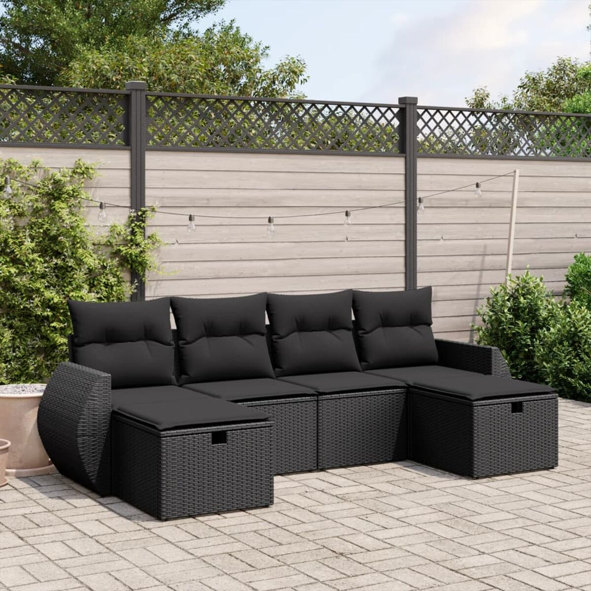 Schwarzes Outdoor-Ecksofa aus Korbgeflecht mit schwarzen Kissen auf einer Terrasse. Lichter sind oben gespannt.
