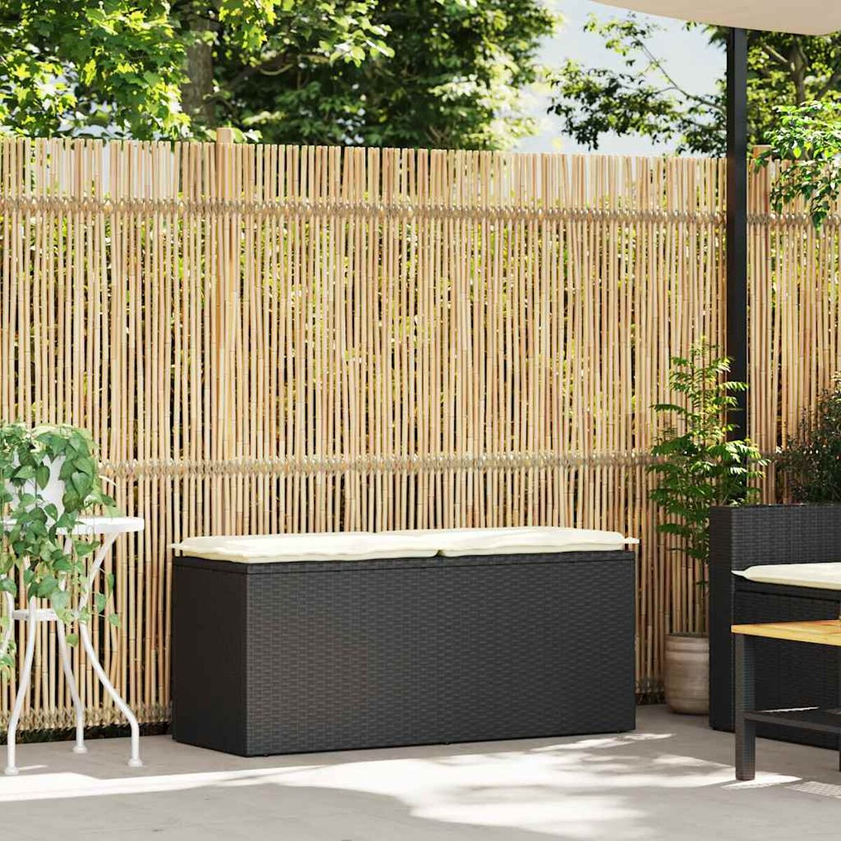 Contenitore nero con coperchio beige, dietro una recinzione di bambù su un patio. Piante di lato.