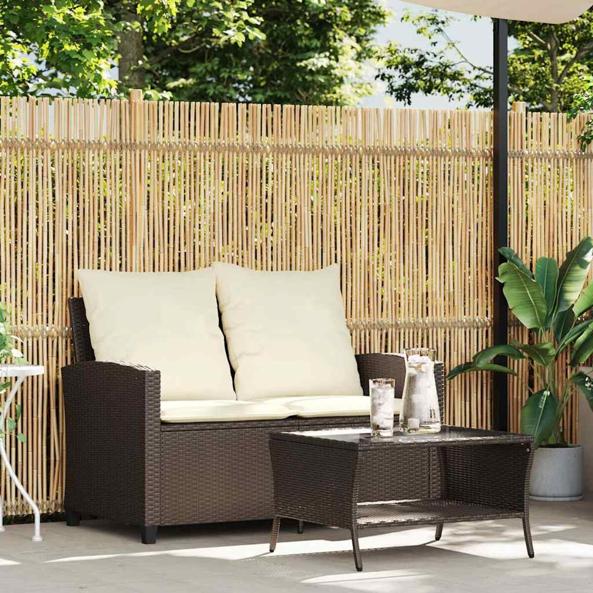 Mobili da patio. Divano in vimini marrone con due cuscini beige, tavolo e piante, recinzione in bambù sul retro.