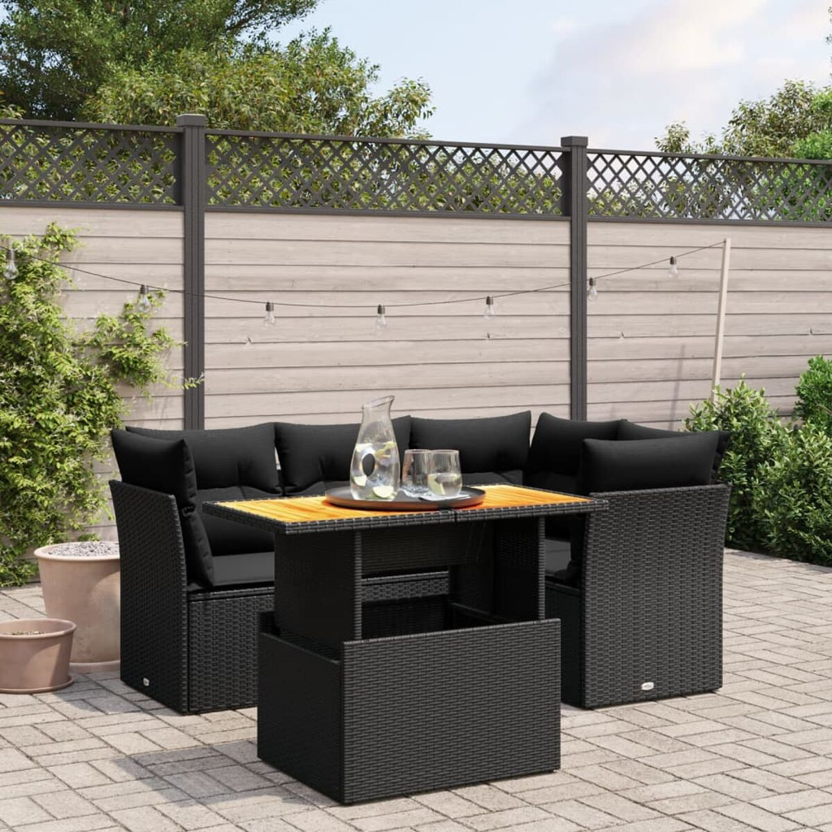 Terrassenset im Freien mit schwarzen Kissen, Tisch und Stühlen auf einer Backsteinterrasse. Ein Zaun und Baum im Hintergrund.