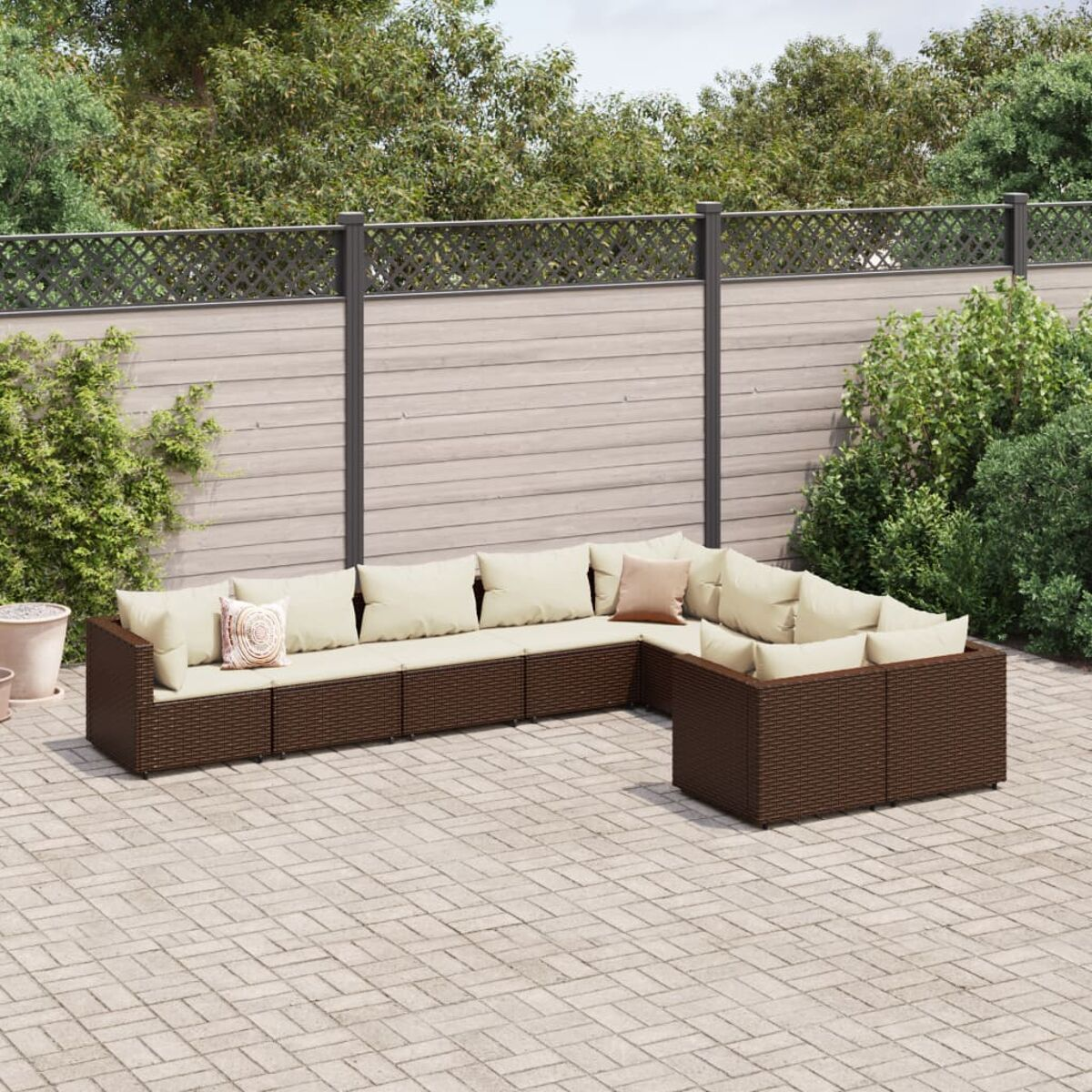 L-förmiges braunes Rattan-Gartensofa mit beigen Kissen, auf einer Steinveranda. Grünes Laub im Hintergrund.
