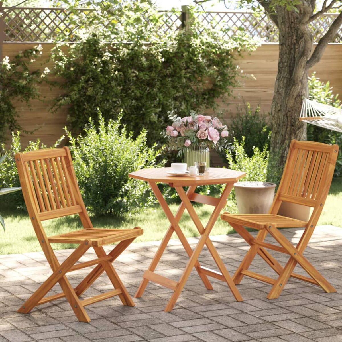 Holztisch und Stühle auf einer Terrasse. Auf dem Tisch steht eine Blumenvase. Grüne Büsche.