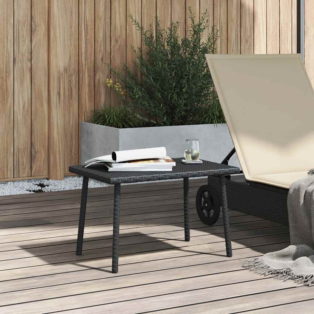 Ein kleiner, schwarzer Korbtisch mit einem Buch, Glas und einer Terrasse mit Liegestuhl im Hintergrund.