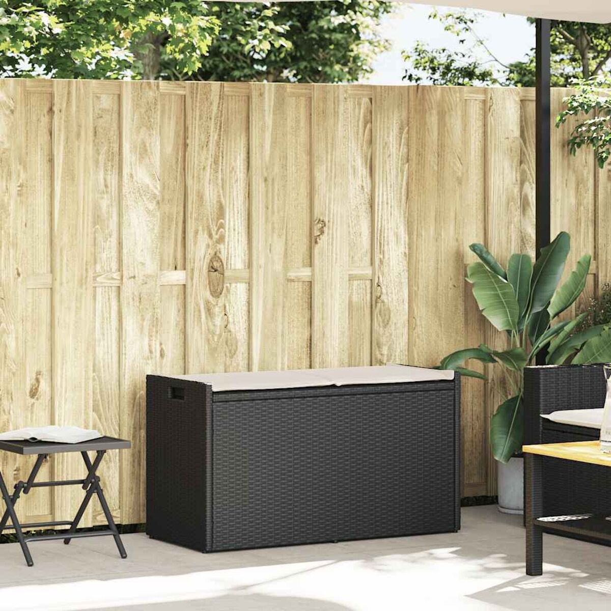 Contenitore portaoggetti da esterno in rattan nero con cuscino beige sopra. Il contenitore è vicino a una recinzione.