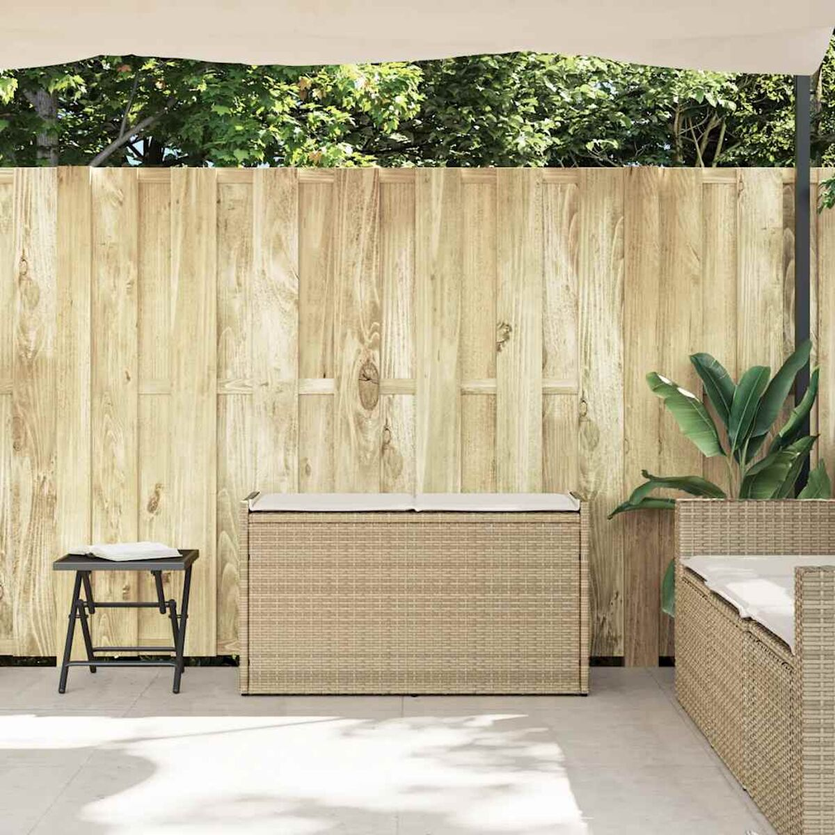 Panca contenitore in rattan beige con cuscino crema. Staccionata in legno e piante sullo sfondo.