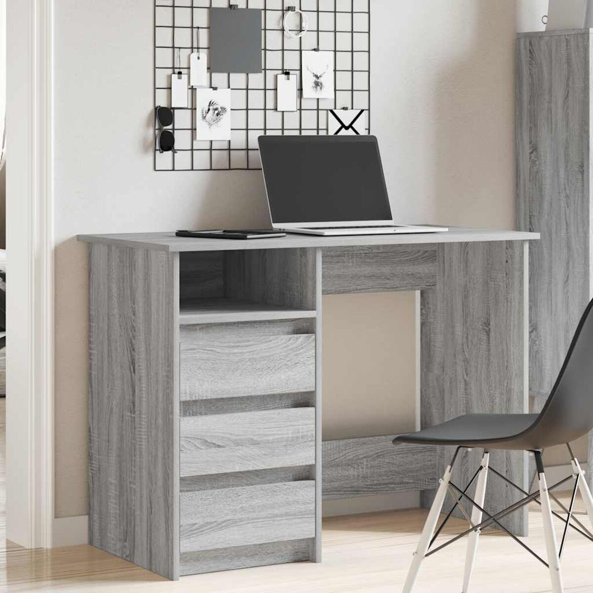 Ein grauer Holzschreibtisch mit Schubladen und einem Laptop, schwarzem Stuhl und Drahtgitter an der Wand.