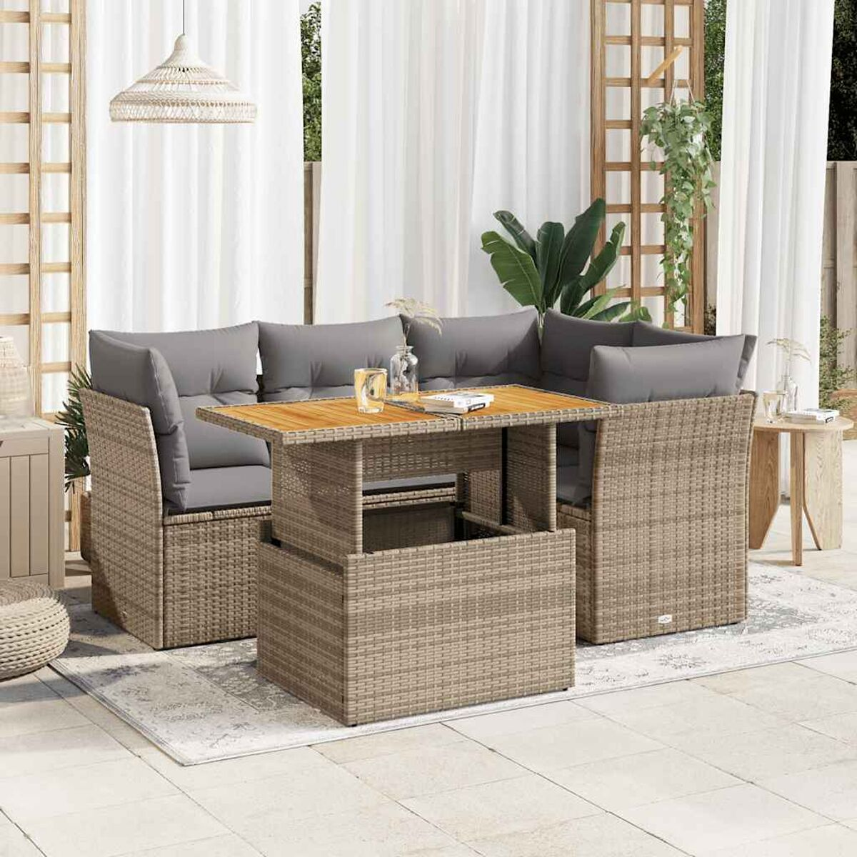 Set da patio in vimini con tavolo sollevabile. Cuscini grigi, tende e piante sullo sfondo.