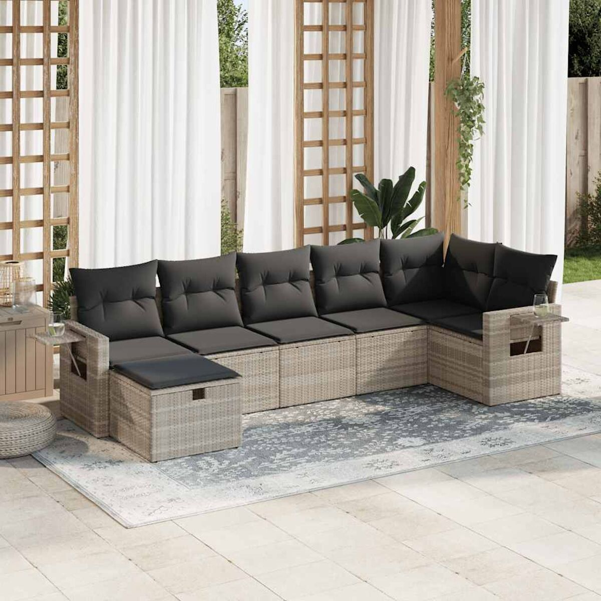Geflochtenes Outdoor-Ecksofa mit grauen Kissen auf einem Teppich. Pflanzen und Zierwand dahinter.