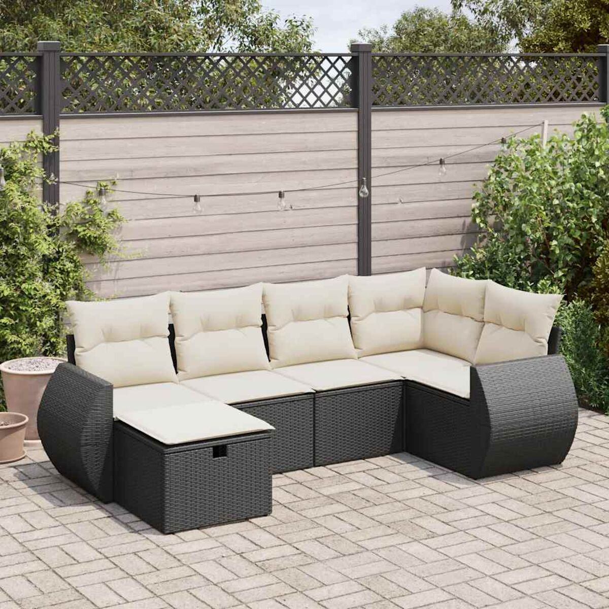 Divano in rattan nero a forma di U con cuscini bianco sporco su un patio in mattoni. Recinzione e piante sullo sfondo.