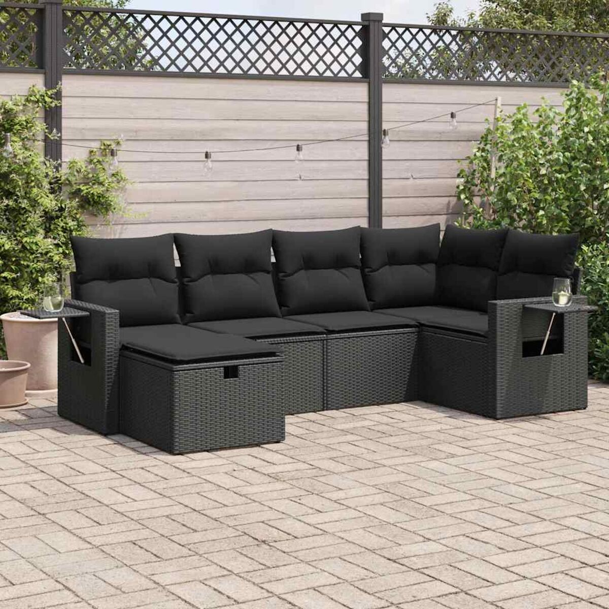Un set per patio esterno con divani in rattan grigio, cuscini neri e un patio in mattoni. Recinzione e piante sullo sfondo.