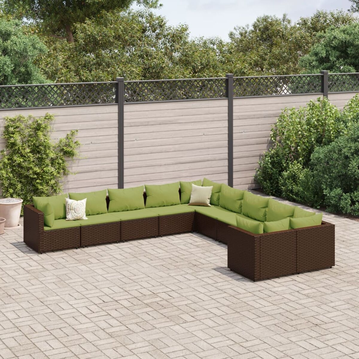 Braunes Outdoor-Ecksofa mit grünen Kissen auf einer Backsteinterrasse, vor einem Zaun und Büschen.