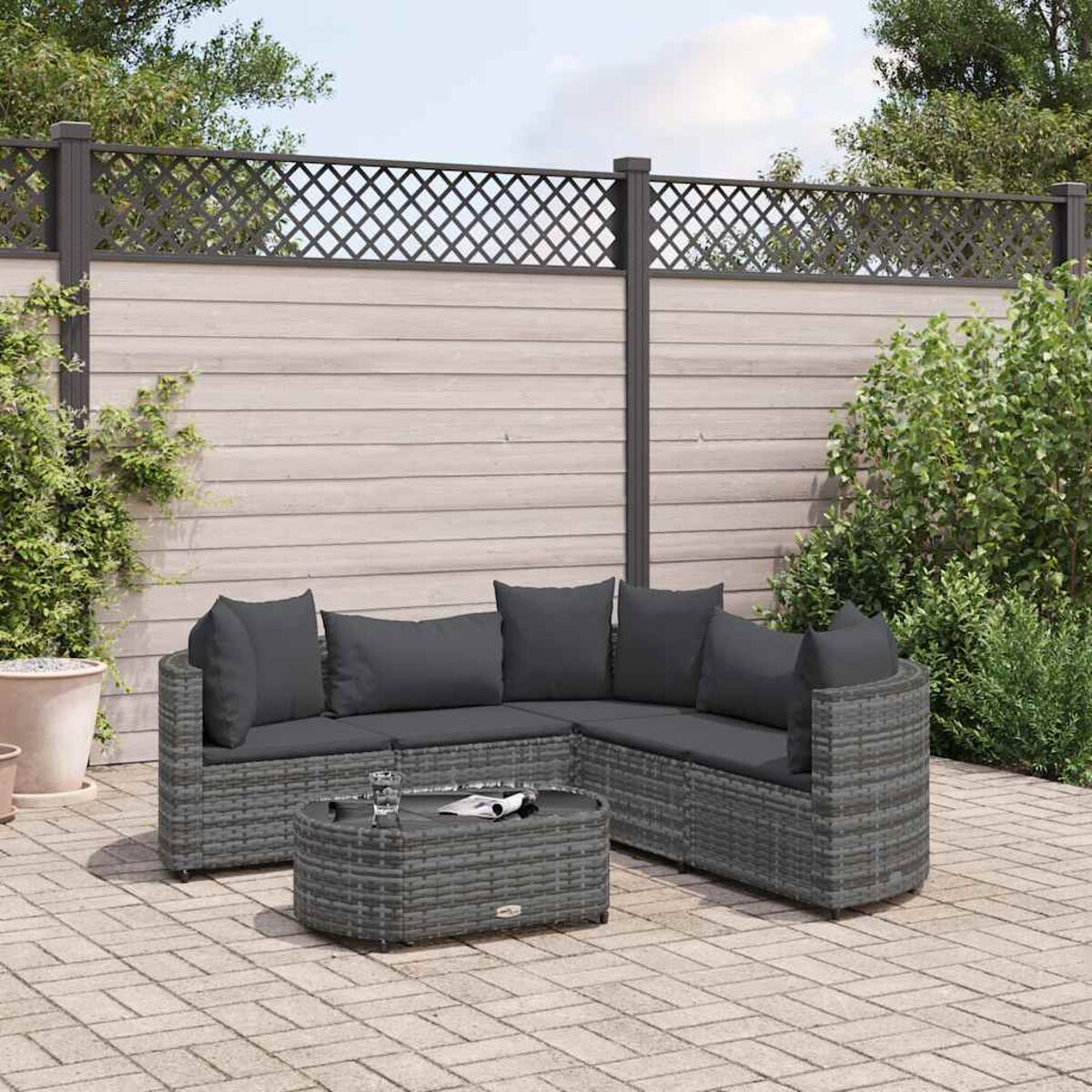 Divano angolare e tavolino in rattan grigio su un patio in pietra, con recinzione e piante sullo sfondo.