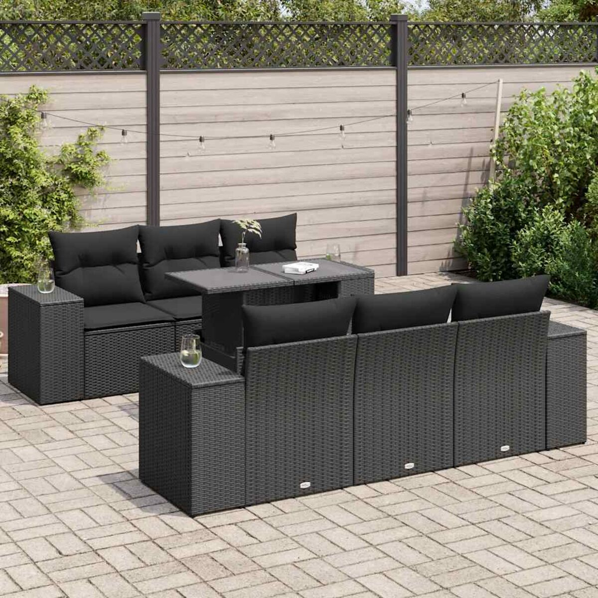 Set di mobili da patio con cuscini neri e tavolo grigio. Lo sfondo mostra recinzione e vegetazione.