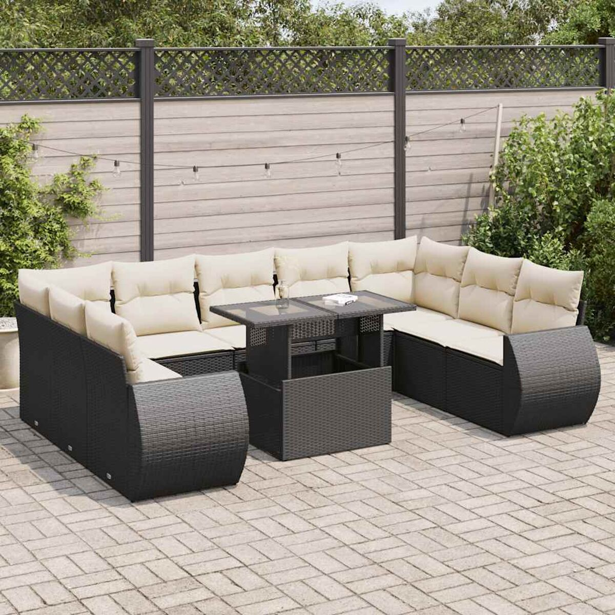 Set divano in vimini nero con cuscini beige, tavolo. All'aperto, su area pavimentata, sfondo con recinzione e piante.