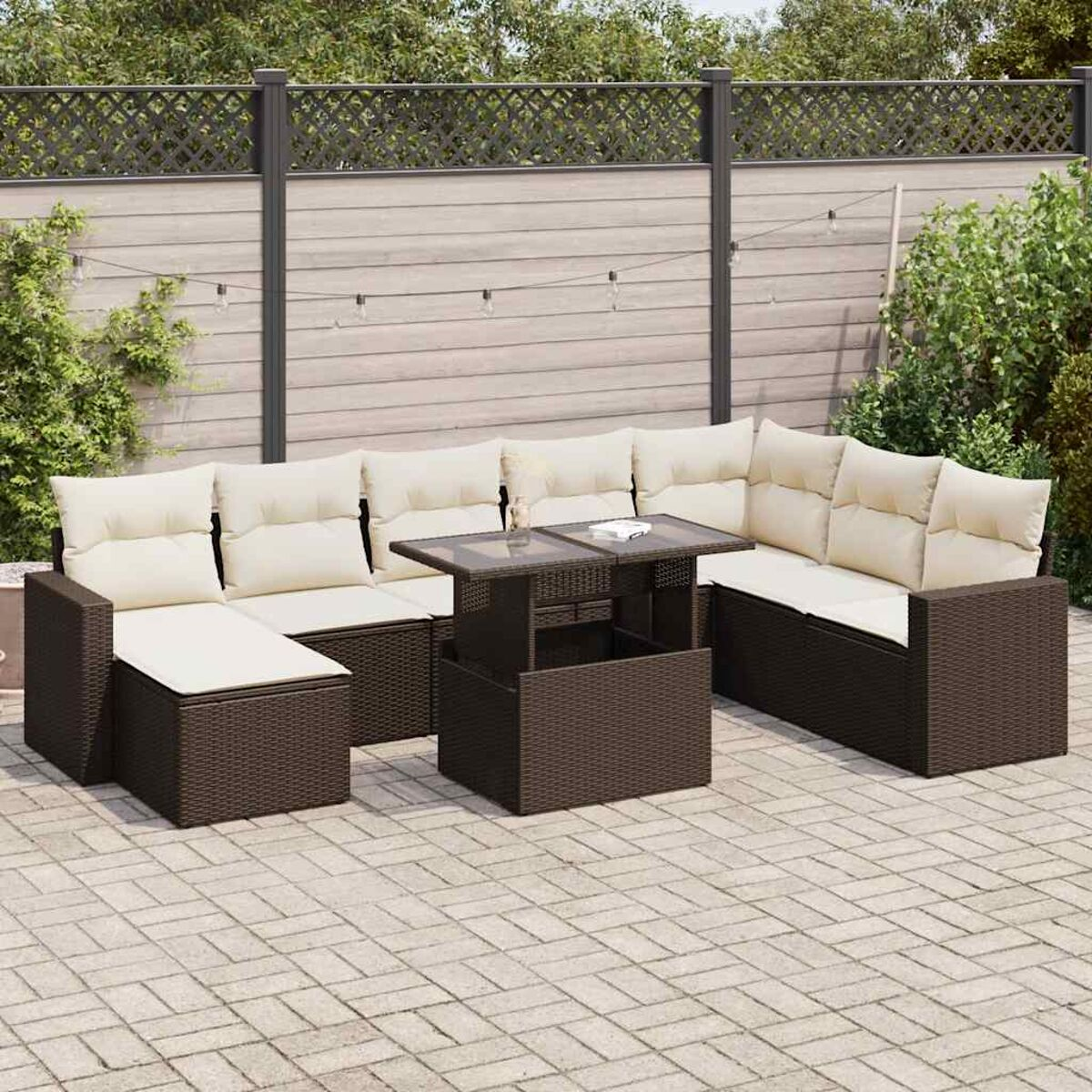Ein Terrassenset mit braunen Korbmöbeln, einem Tisch und cremefarbenen Kissen. Der Hintergrund zeigt einen Zaun und Pflanzen.