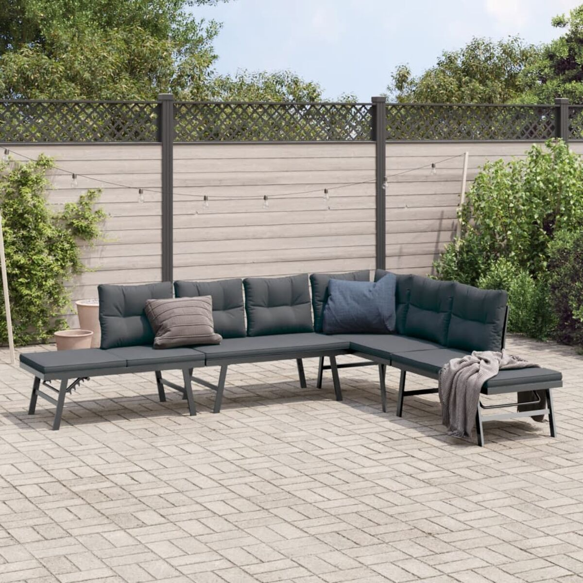 Ecksofa im Freien mit grauen Kissen auf einer Ziegelterrasse, Zaun im Hintergrund.