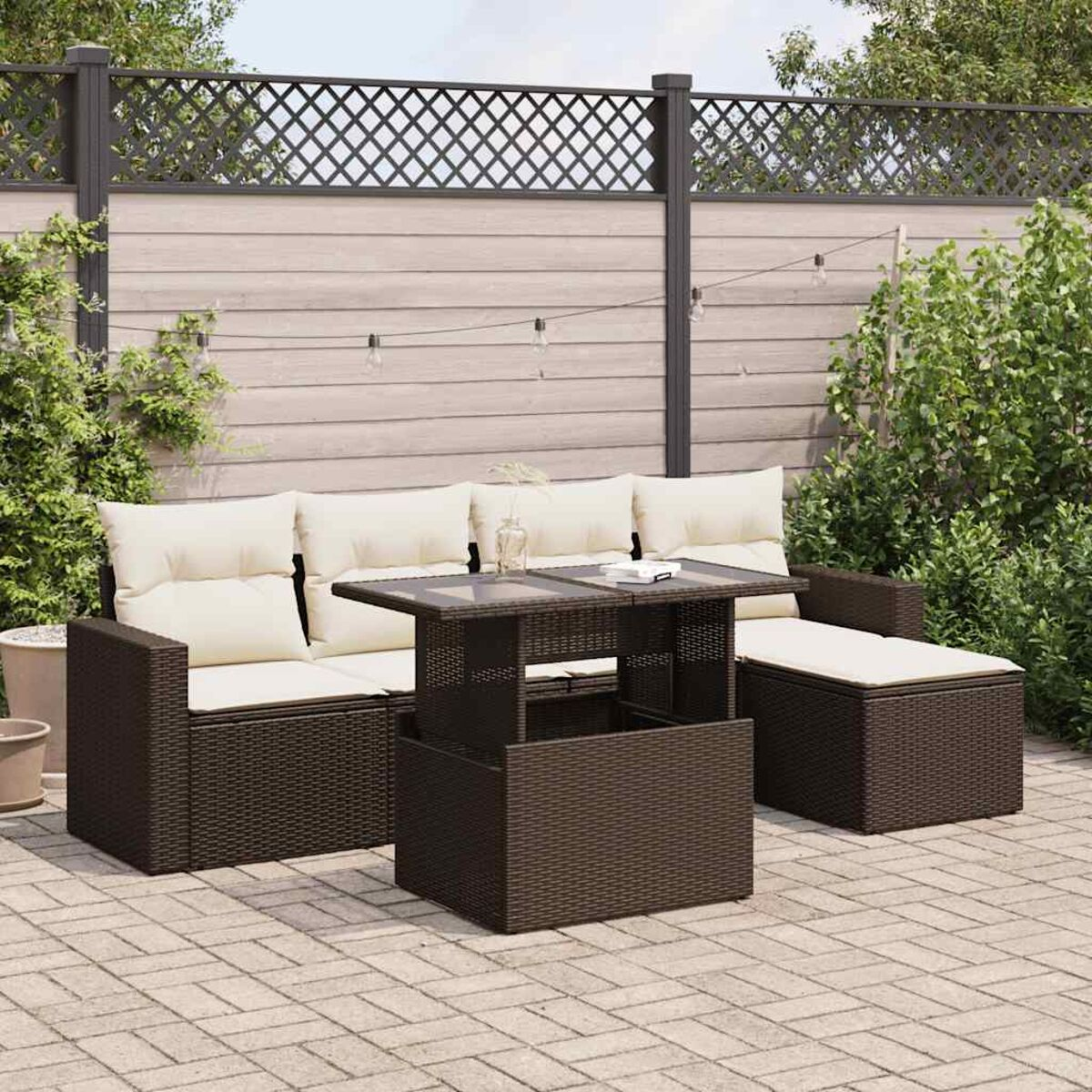 Braunes Outdoor-Set aus Korbgeflecht mit beigefarbenen Kissen und Tisch auf einer Backstein-Terrasse. Holzzaun im Hintergrund.
