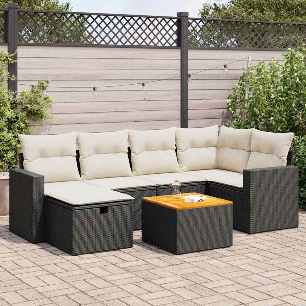 Schwarzes Rattan-Ecksofa mit cremefarbenen Kissen, Holztischplatte. Ziegelterrasse, Zaun und Pflanzen im Hintergrund.