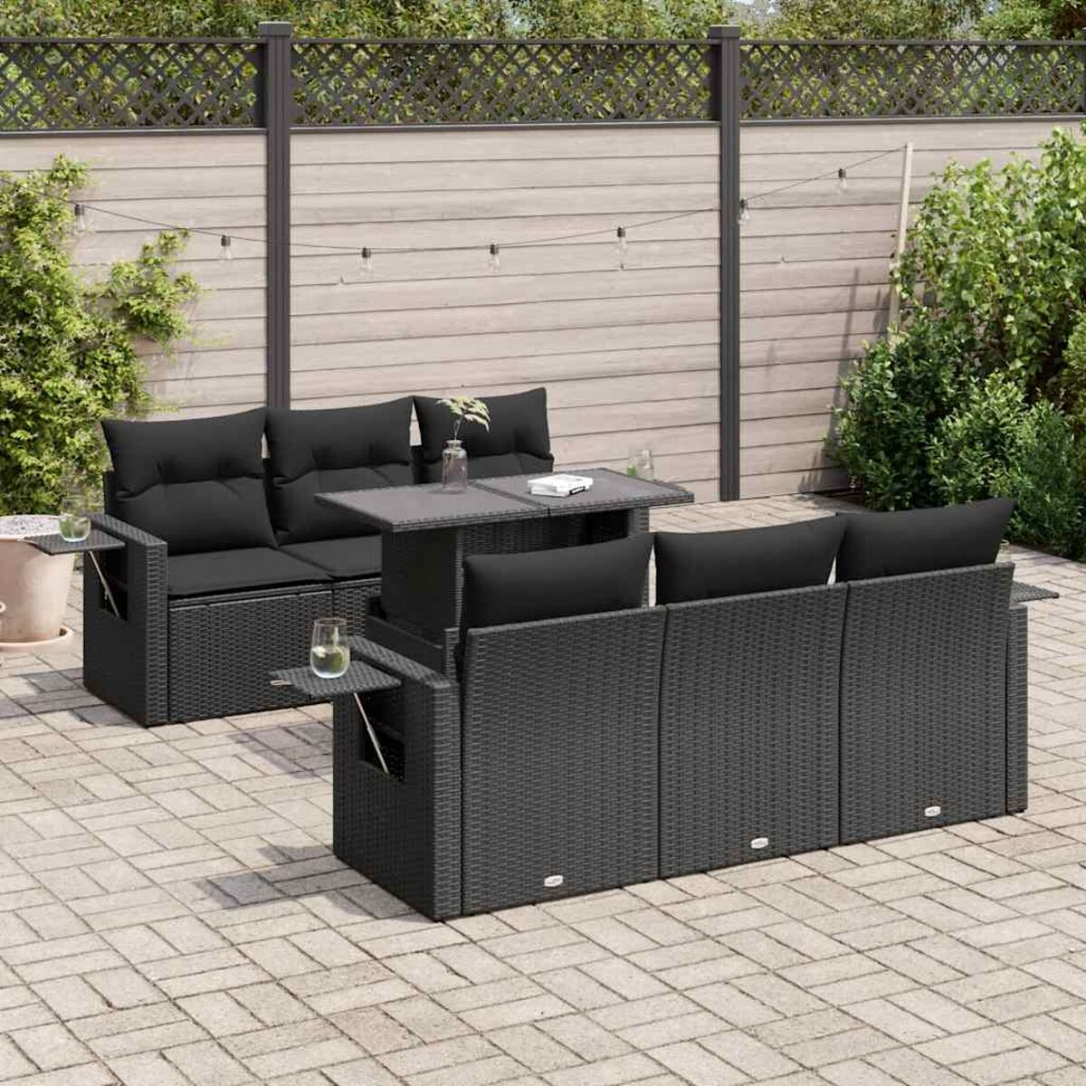 Set divano in rattan nero con tavolo su un patio in pietra, con piante e recinzione sullo sfondo.