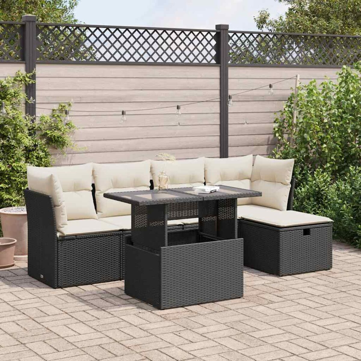 Divano componibile in rattan nero per esterni con cuscini bianchi, un tavolo di vetro. Sfondo con recinzione e piante.