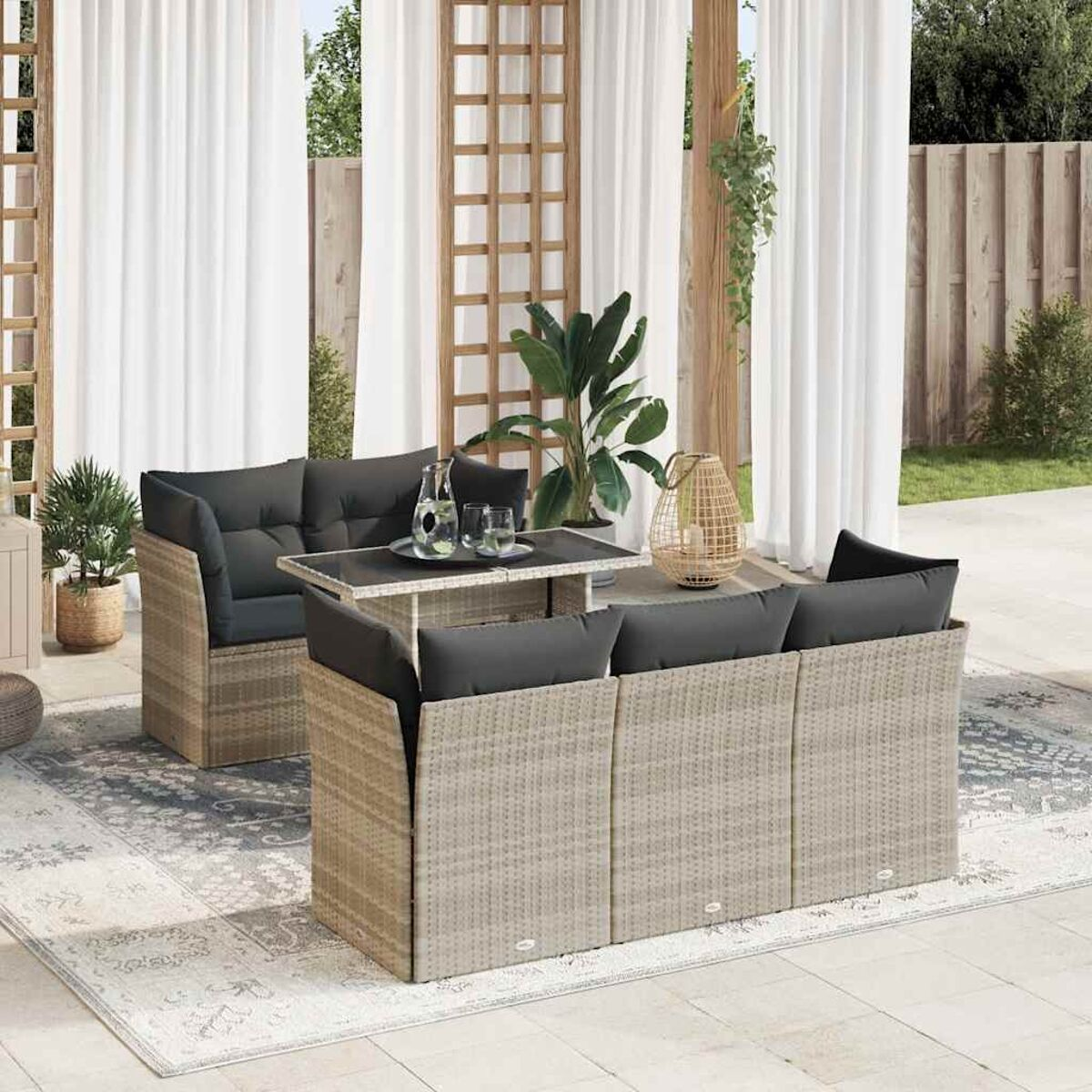 Set in rattan per esterni con cuscini, tavolo, piante e decorazioni. Scena all'aperto con tende bianche.