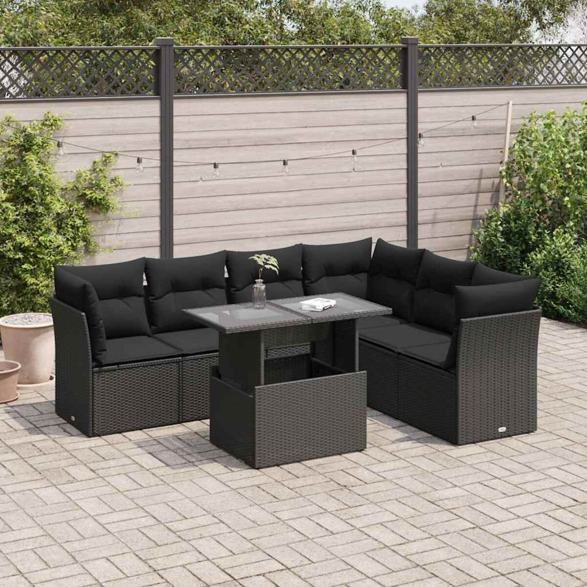 Divano angolare da esterno in vimini nero con tavolo con piano in vetro su un patio pavimentato, con una recinzione sullo sfondo.