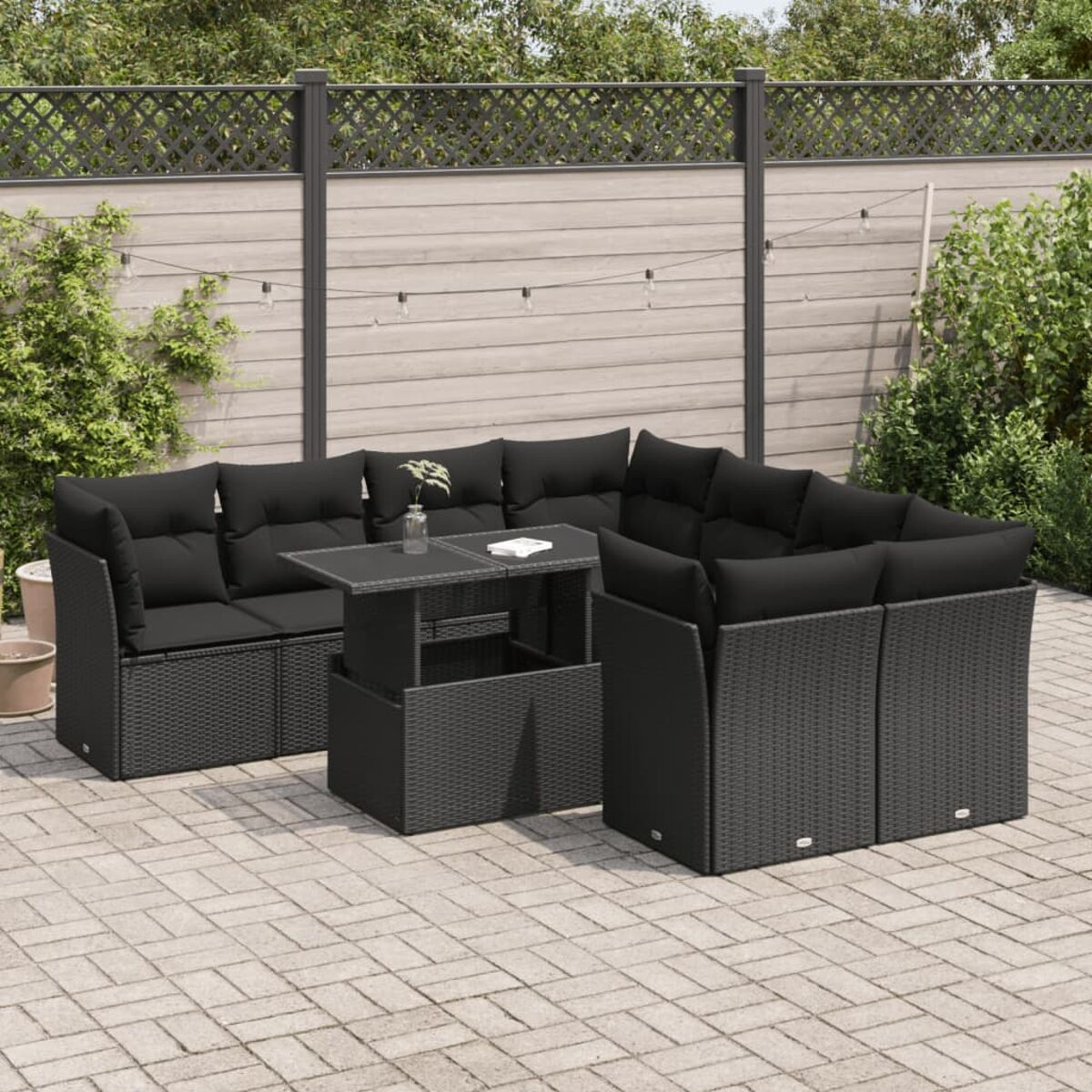 Set divani in vimini nero da esterno con tavolo e cuscini neri su un patio, con piante e recinzione.