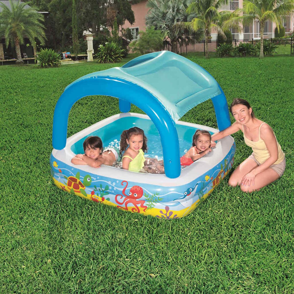 Une femme et trois enfants dans une piscine gonflable bleue et blanche sur une pelouse.