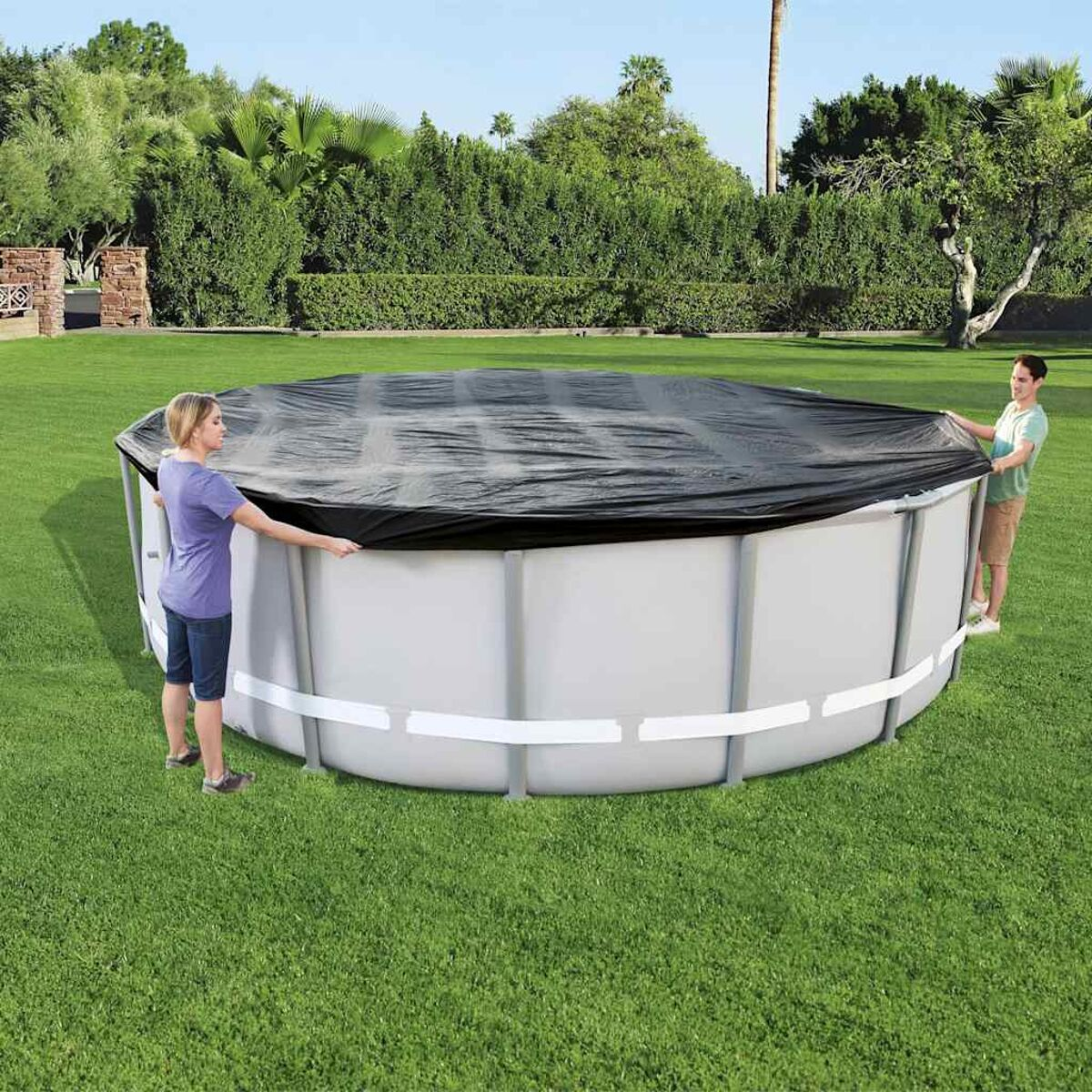 Zwei Personen bedecken einen runden Pool mit einer schwarzen Abdeckung. Der Pool ist silbern und auf Gras.