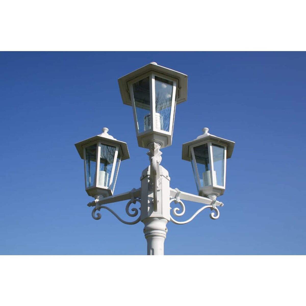 Eine weiße verzierte Straßenlaterne mit drei Lichtern vor blauem Himmel.