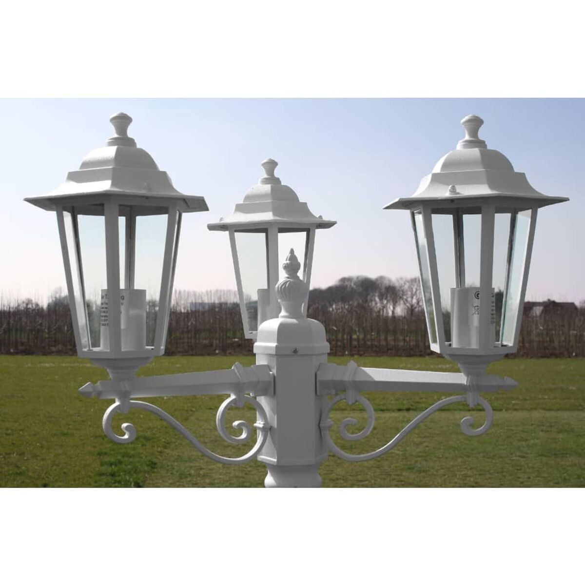 Drei weiße Außenlaternen an einem Pfahl. Der Hintergrund ist eine Wiese und ein blauer Himmel.