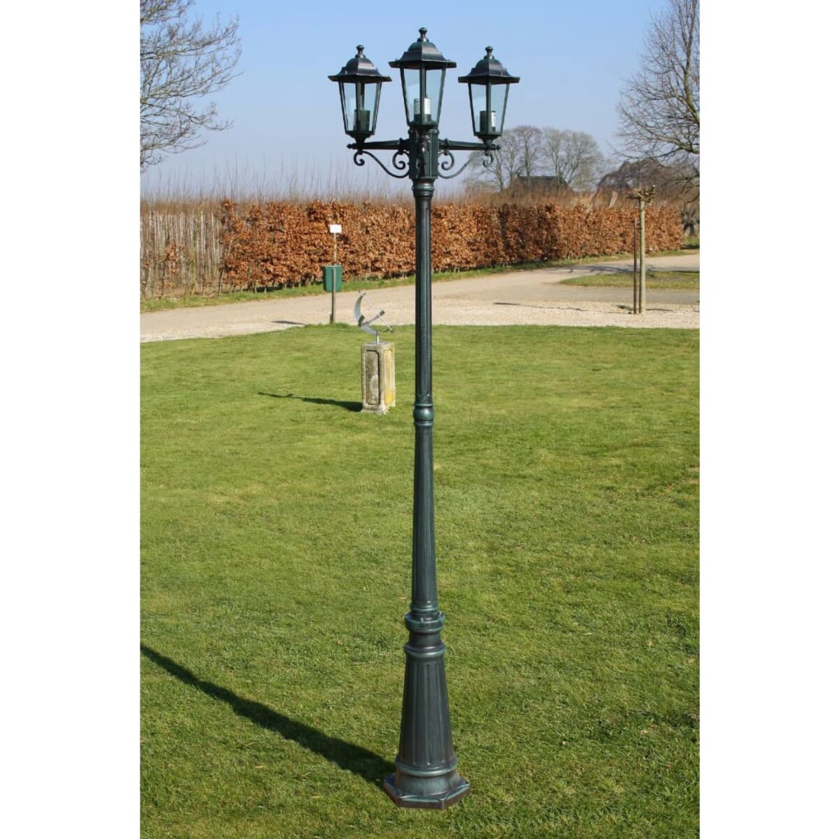 Dreilampige schwarze Straßenlaterne auf Gras. Grünes Gras und Bäume im Hintergrund.