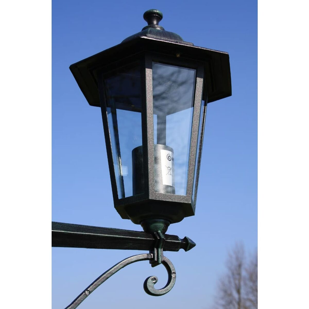 Schwarze Außenlampe mit einem Gehäuse aus klarem Glas. Die Lampe ist an einer Wand mit blauem Himmel montiert.
