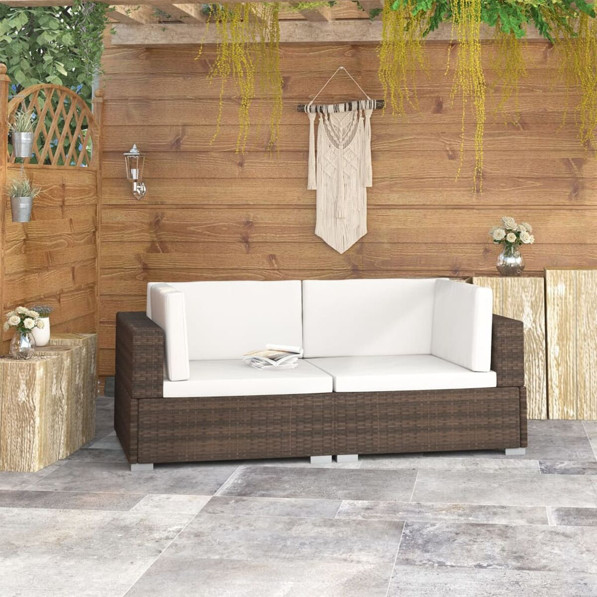 Braunes Korb-Outdoor-Sofa mit weißen Kissen, auf einer Steinterrasse mit Holzwand im Hintergrund.