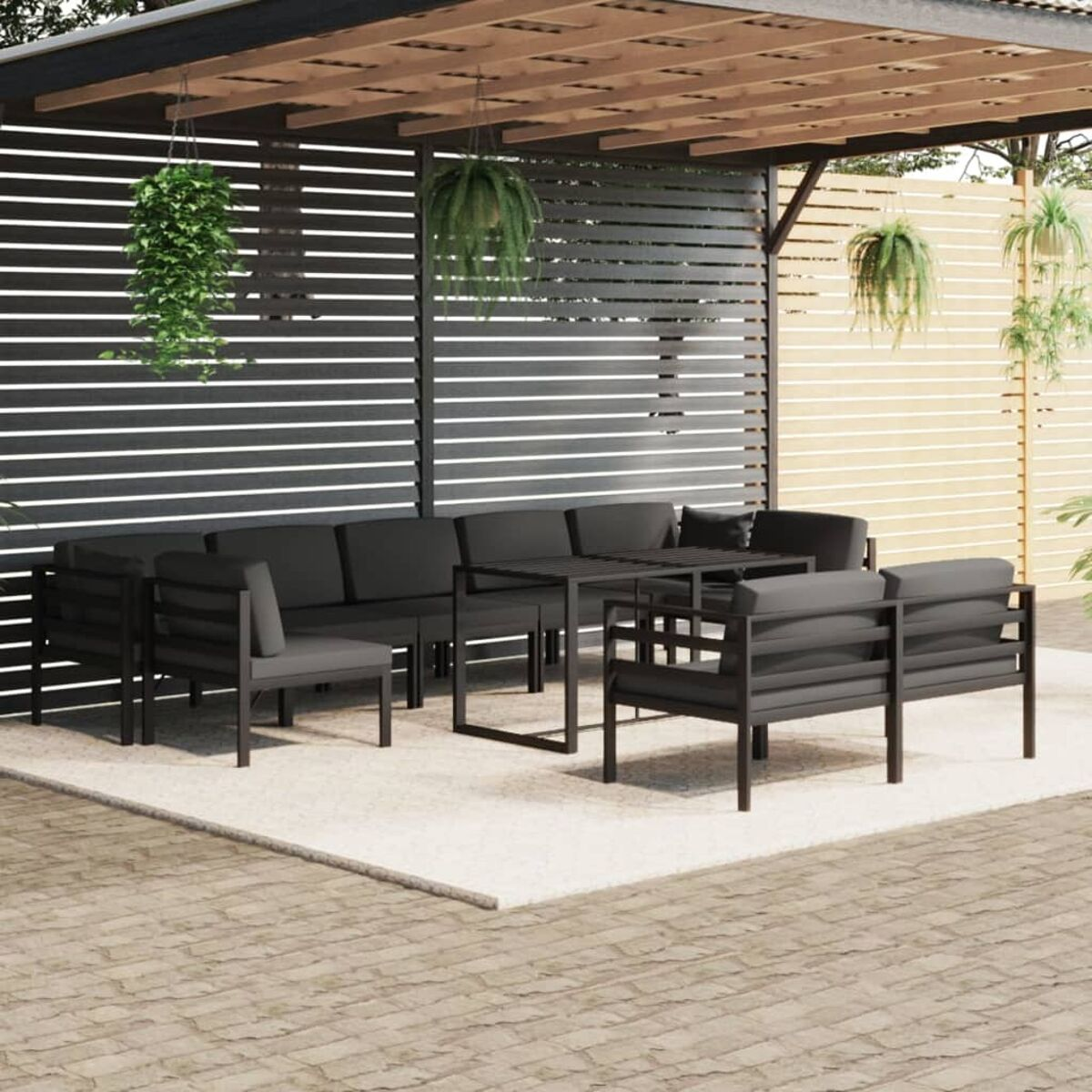 Garten-Sitzgruppe. Schwarzer Rahmen, graue Kissen. Pflanzen hängen von der Pergola. Beiger Teppich auf Ziegelpflaster.