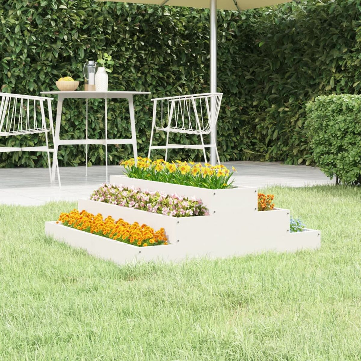 Mehrstufiger weißer Pflanzkübel mit Blumen, grüner Rasen, Büsche im Hintergrund.