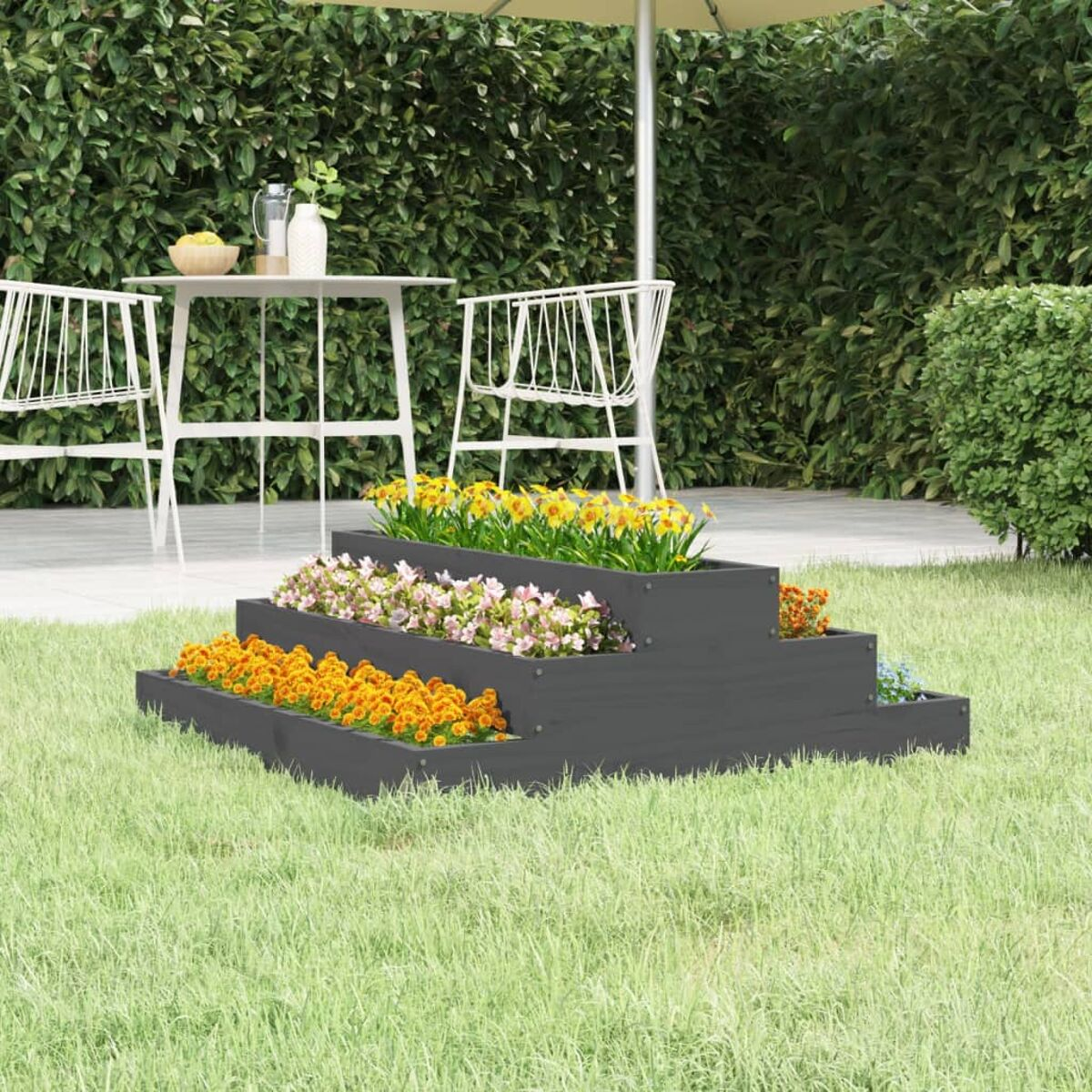 Ein abgestufter, grauer Pflanzkübel mit Blumen steht auf Gras. Im Hintergrund ein Tisch und Stühle.