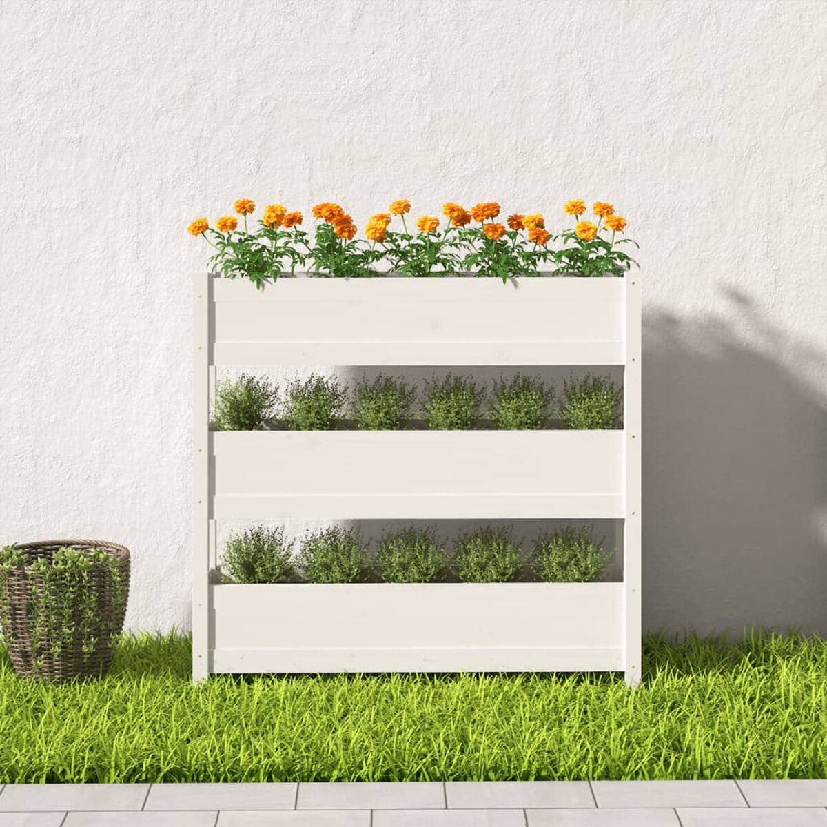 Weißer vertikaler Holzpflanzer mit Pflanzen, orangefarbenen Blumen, Gras und einem Weidenkorb. Sonniger Tag.