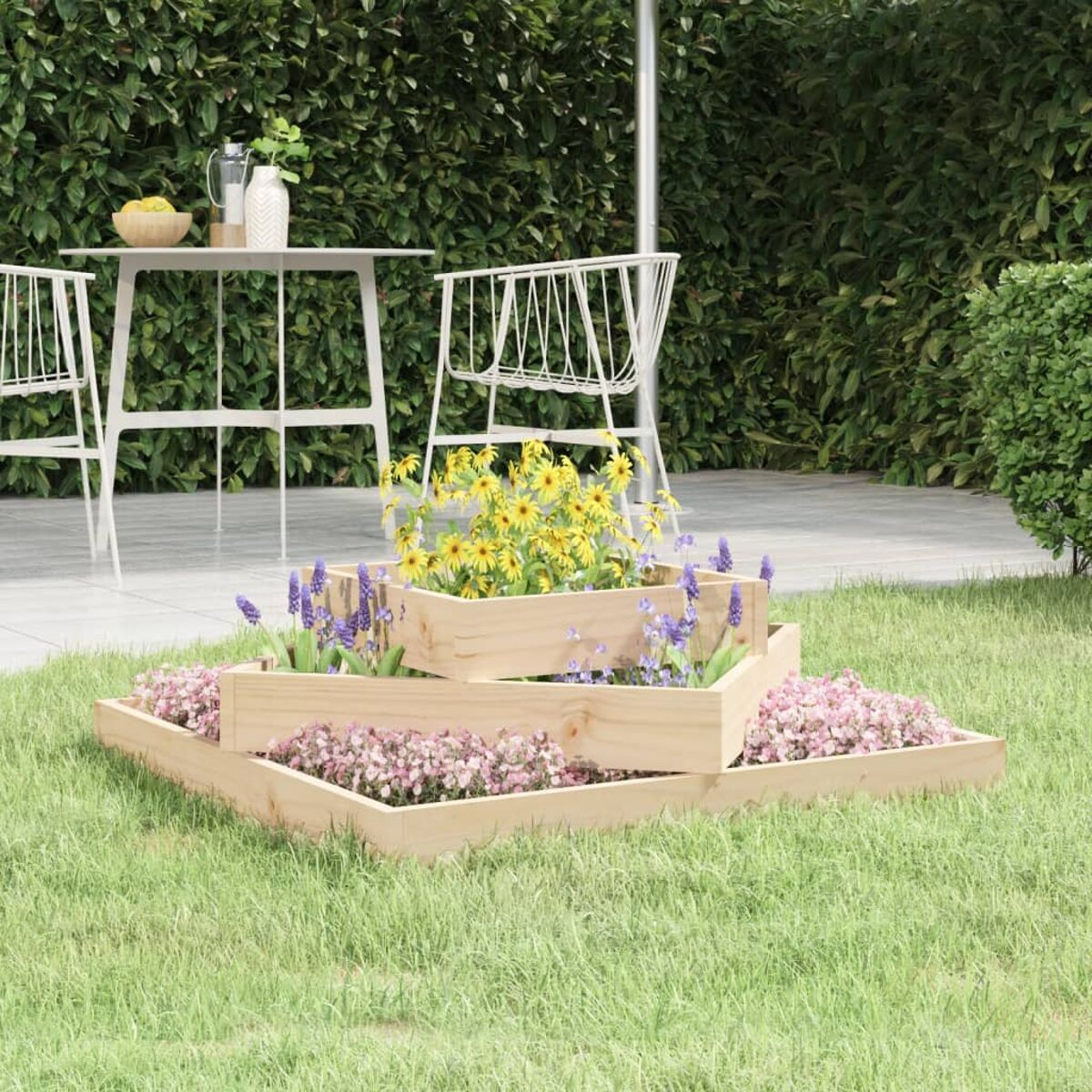 Ein gestufter Holzpflanzer mit Blumen steht im Gras, im Hintergrund ein Tisch und Stühle.