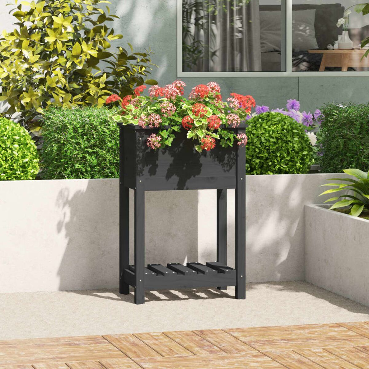 Schwarzer Pflanzkasten mit bunten Blumen. Er steht auf einer Terrasse, umgeben von Grün und einer hellen Wand.
