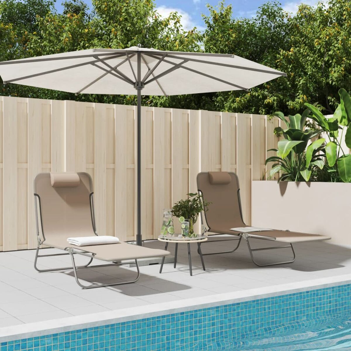Zwei Liegestühle, Tisch mit Getränken und Sonnenschirm an einem Pool. Grüne Pflanzen und ein Zaun.