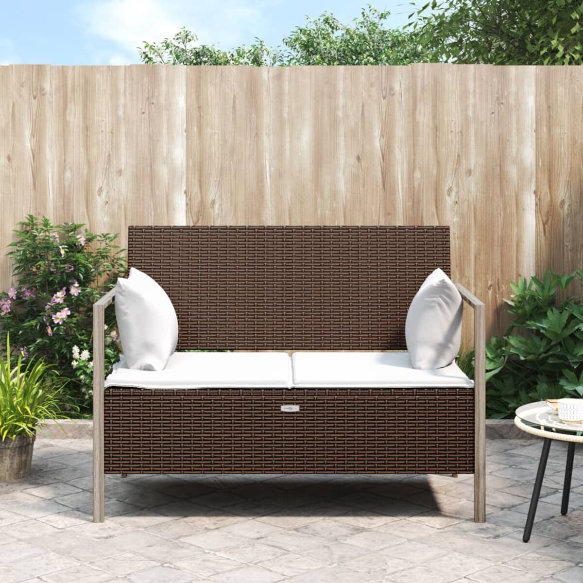 Braunes Outdoor-Liebesitz aus Korbgeflecht mit weißen Kissen, in einem Garten mit Holzzaun.