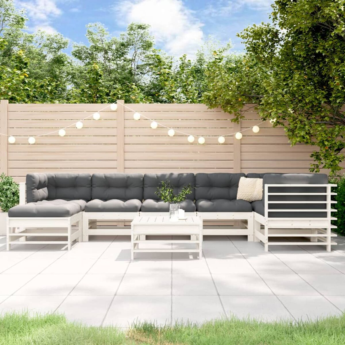 Outdoor-Sitzgruppe mit grauen Kissen, Holztisch und Holzzaun. Lichterketten sind angebracht.